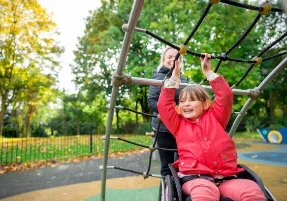 A cheerful low angle medium close-up of a young girl who is a wheelchair user playing in the park on the climbing frame with her mother. They're in a playground ina. public park in Newcastle upon Tyne in the North East of England